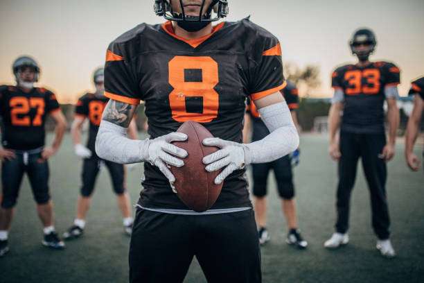 Player in jersey #8 grips football with white football gloves, teammates in background.