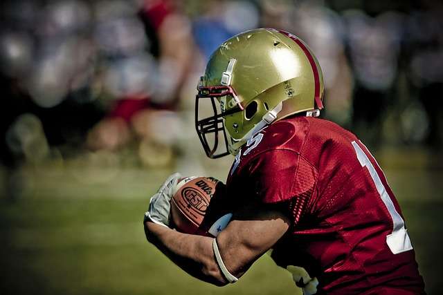 Football player in red jersey and gold helmet catching a ball, wearing football gloves.