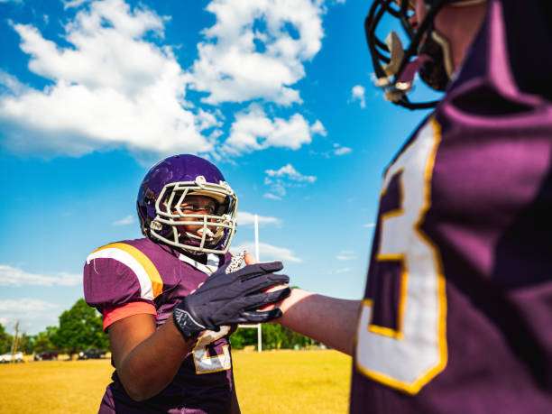 Two young football players in purple jerseys and football gloves shaking hands on field.