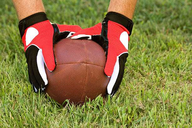 Hands wearing red and white football gloves placing a football on green grass.