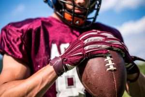 Football player in maroon jersey wearing matching football gloves, holding a ball outdoors.