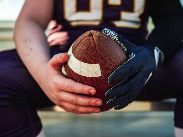 Close-up of football player's hands gripping a football, one wearing black football gloves.